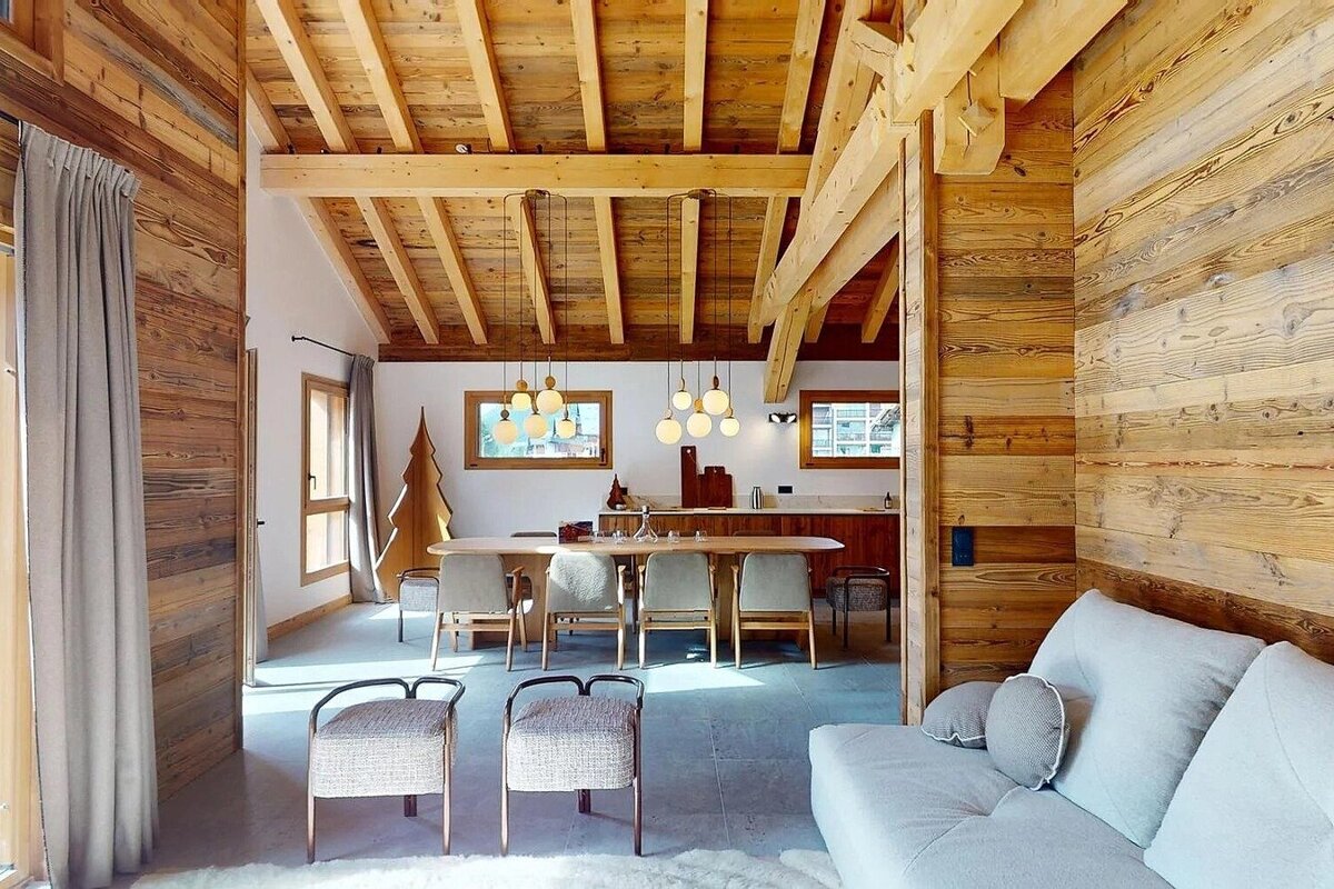 Rustic wooden chalet interior featuring exposed ceiling beams, a dining table with pendant lights, and a cozy sofa area.