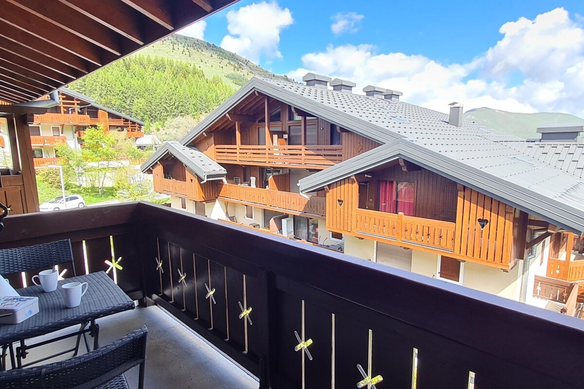 Balcony view of a mountain village with charming wooden chalets against lush green mountains and a blue sky. A table with cups is on the balcony.