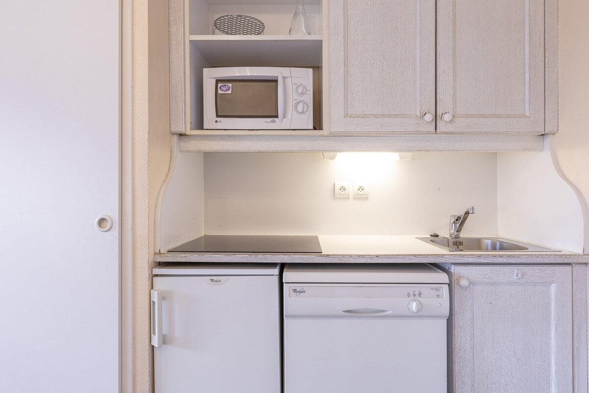 A compact, light-toned kitchenette featuring a microwave, cooktop, sink, refrigerator, and dishwasher within wooden cabinets.