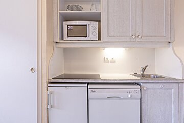A compact, light-toned kitchenette featuring a microwave, cooktop, sink, refrigerator, and dishwasher within wooden cabinets.