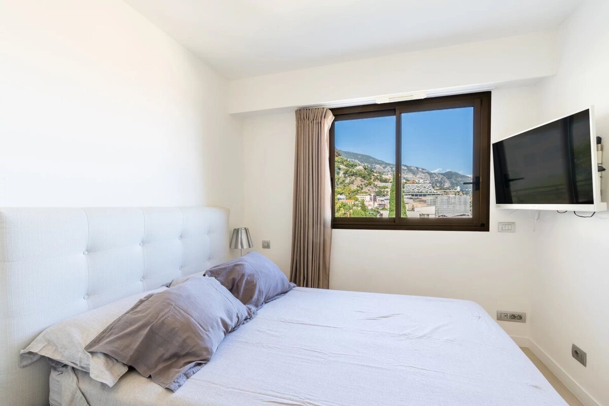 A bright, modern bedroom featuring a large white tufted bed, a wall-mounted TV, and a window with a scenic view of a hillside town.