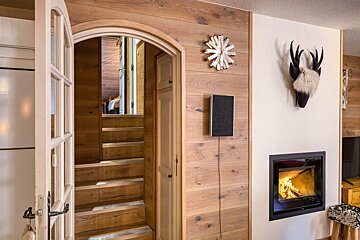 A rustic interior features wood-paneled walls, an arched doorway leading to stairs, a cozy fireplace, and a deer head decoration.