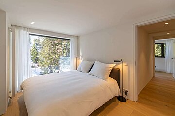 Modern bedroom with a large window showcasing a snowy forest. Features a white bed, light wood floors, and minimalist lighting, offering a serene escape.
