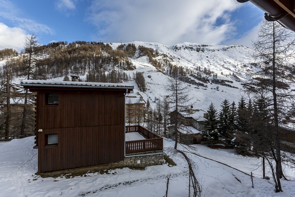 Wooden chalets nestled in a snowy alpine valley with pine trees and snow-covered mountains under a blue sky.