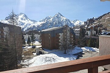 Sunny view from a wooden balcony of a mountain resort with snow-capped peaks, pine trees, and apartment buildings.