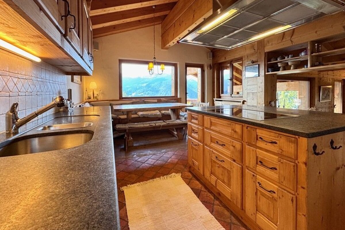 A cozy, rustic wooden kitchen in a mountain chalet, featuring warm lighting, a central island, and scenic views from the window.