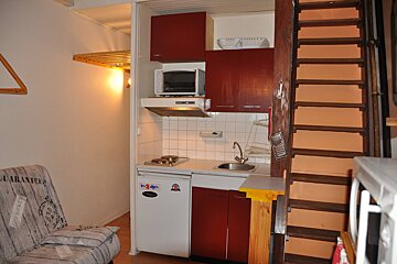 A cozy studio featuring a compact kitchenette with red cabinets, microwave, stovetop, and fridge, alongside a sofa and wooden loft ladder.