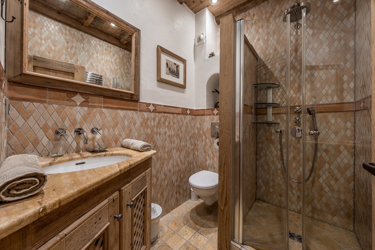 Cozy, rustic bathroom with diamond-patterned terracotta tiles, wooden vanity, mirror, toilet, and glass shower cabin in warm chalet style.