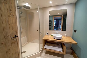 A modern bathroom featuring a glass shower, wooden vanity with a vessel sink, and a mirror reflecting an adjacent room, against light blue walls.
