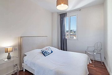 A bright, minimalist bedroom features white walls, a bed with white bedding, a clear nightstand with lamp, window, and a ghost chair on wooden floors.