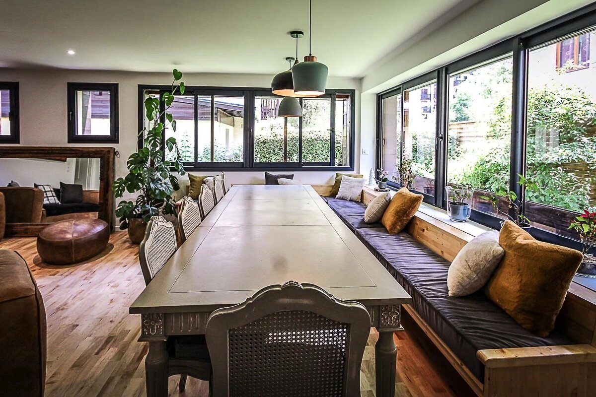 A long dining table with traditional chairs and modern bench seating along large windows overlooking a green garden.