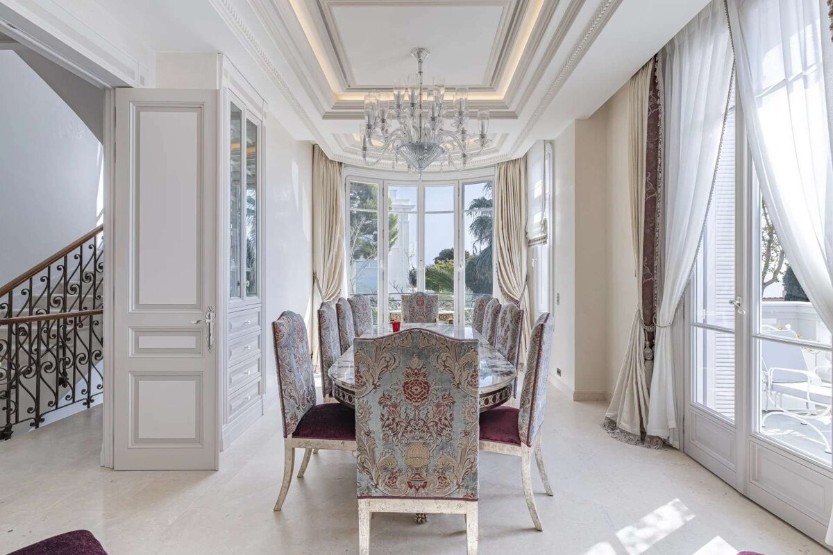 Luxurious dining room with an ornate table, patterned chairs, chandelier, and panoramic windows offering an outdoor view. A staircase is to the left.