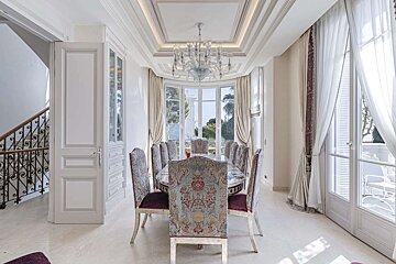 Luxurious dining room with an ornate table, patterned chairs, chandelier, and panoramic windows offering an outdoor view. A staircase is to the left.