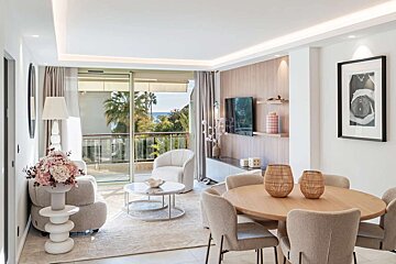 Modern living/dining room with a balcony overlooking palm trees and the sea. Features contemporary furniture, neutral tones, and bright natural light.