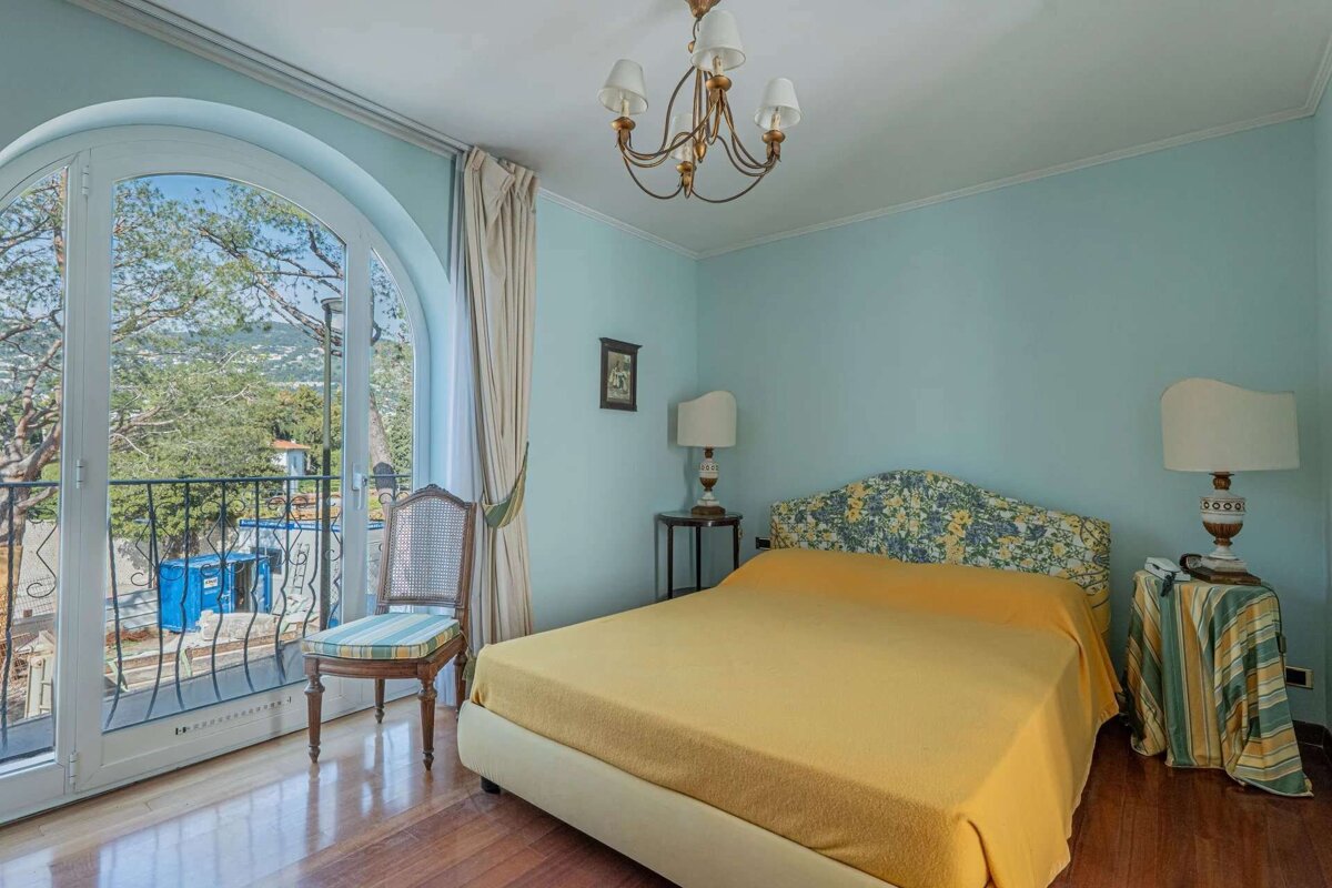 A bright bedroom with light blue walls, a yellow bed, and a floral headboard. A large arched door offers a scenic view. Features a chandelier and wooden floor.