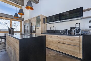 A modern kitchen with light wood cabinets, dark counters, and a stunning snowy mountain view through large windows.