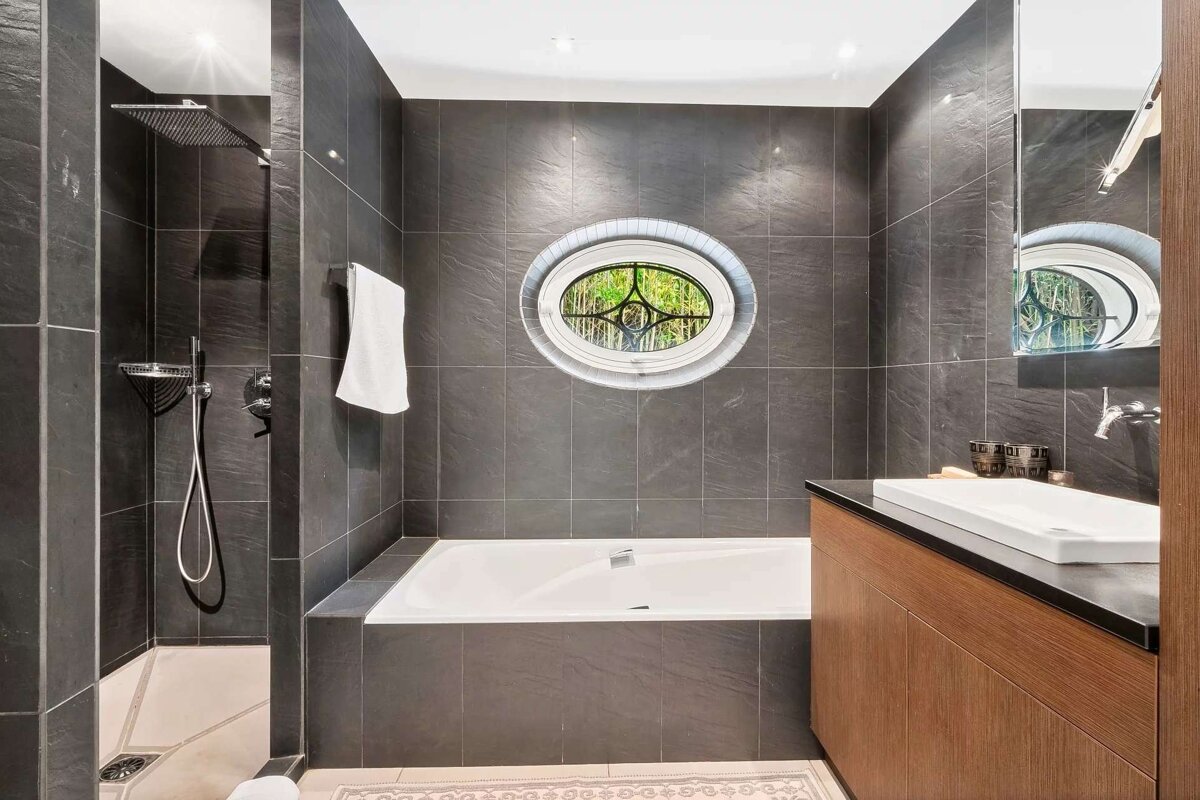 A modern bathroom with dark gray tiled walls, a built-in tub, an oval window, a separate shower, and a wooden vanity with a white sink.