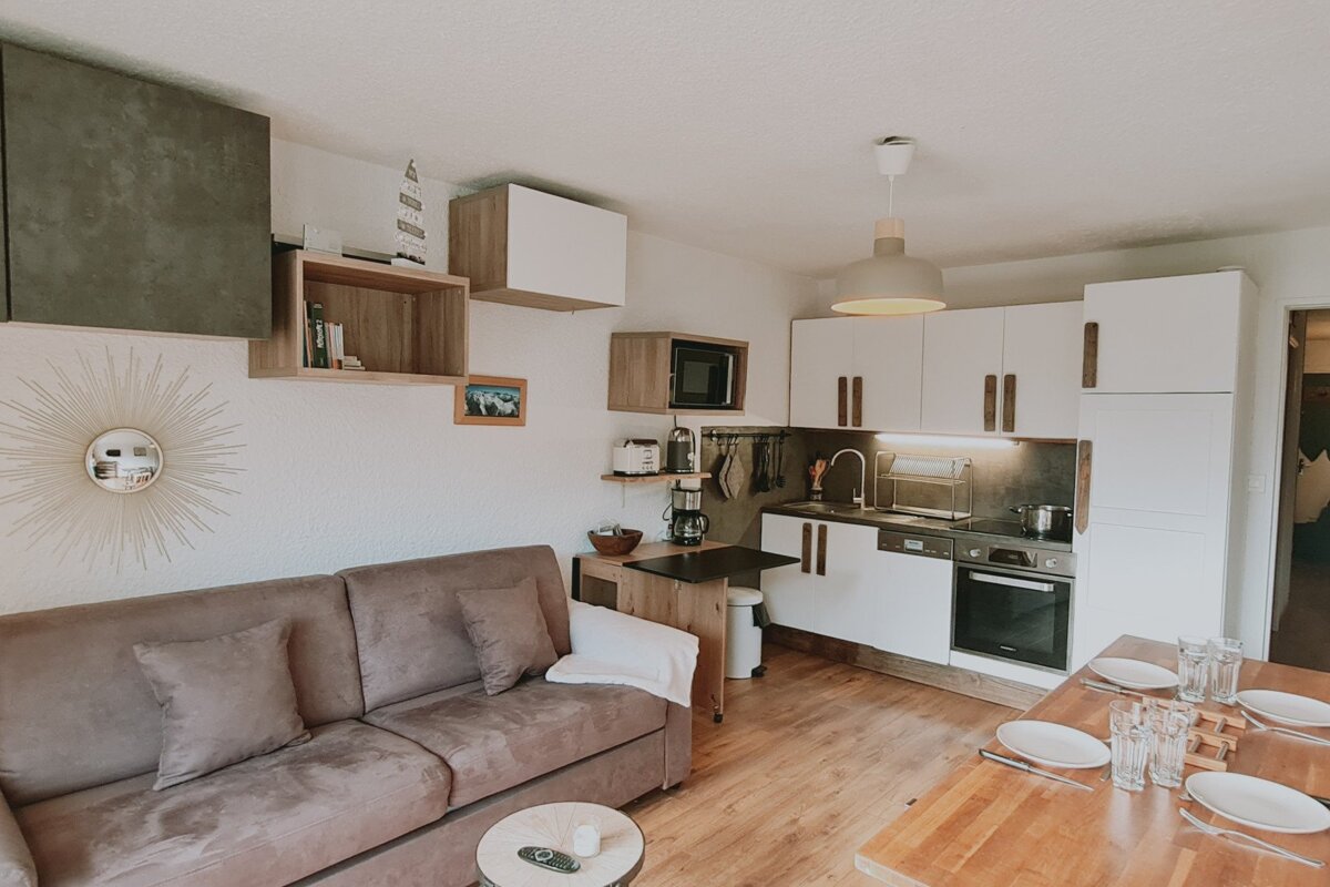 A cozy, modern open-plan space with a brown sofa, compact kitchen, and dining table. White cabinets, wooden floors, and stylish wall decor create a welcoming feel.
