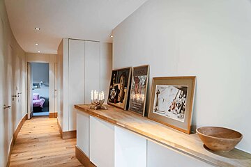 A sleek, light-filled hallway featuring light wood flooring, white built-in storage with art and decor, leading to another room with a purple bed.