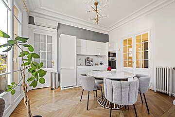 A bright, modern kitchen and dining area with white cabinets, a round table, grey chairs, herringbone wood floors, and large glass doors.