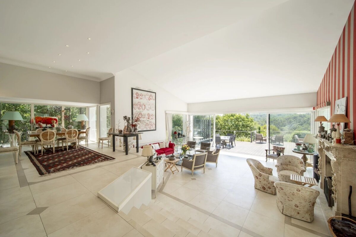 Spacious, bright living/dining room with varied seating, light floors, a red striped accent wall, and large windows opening to a green outdoor deck.