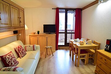 Cozy chalet-style apartment featuring a cream sofa with red pillows, wooden furniture, a dining set, and red curtains leading to a balcony.