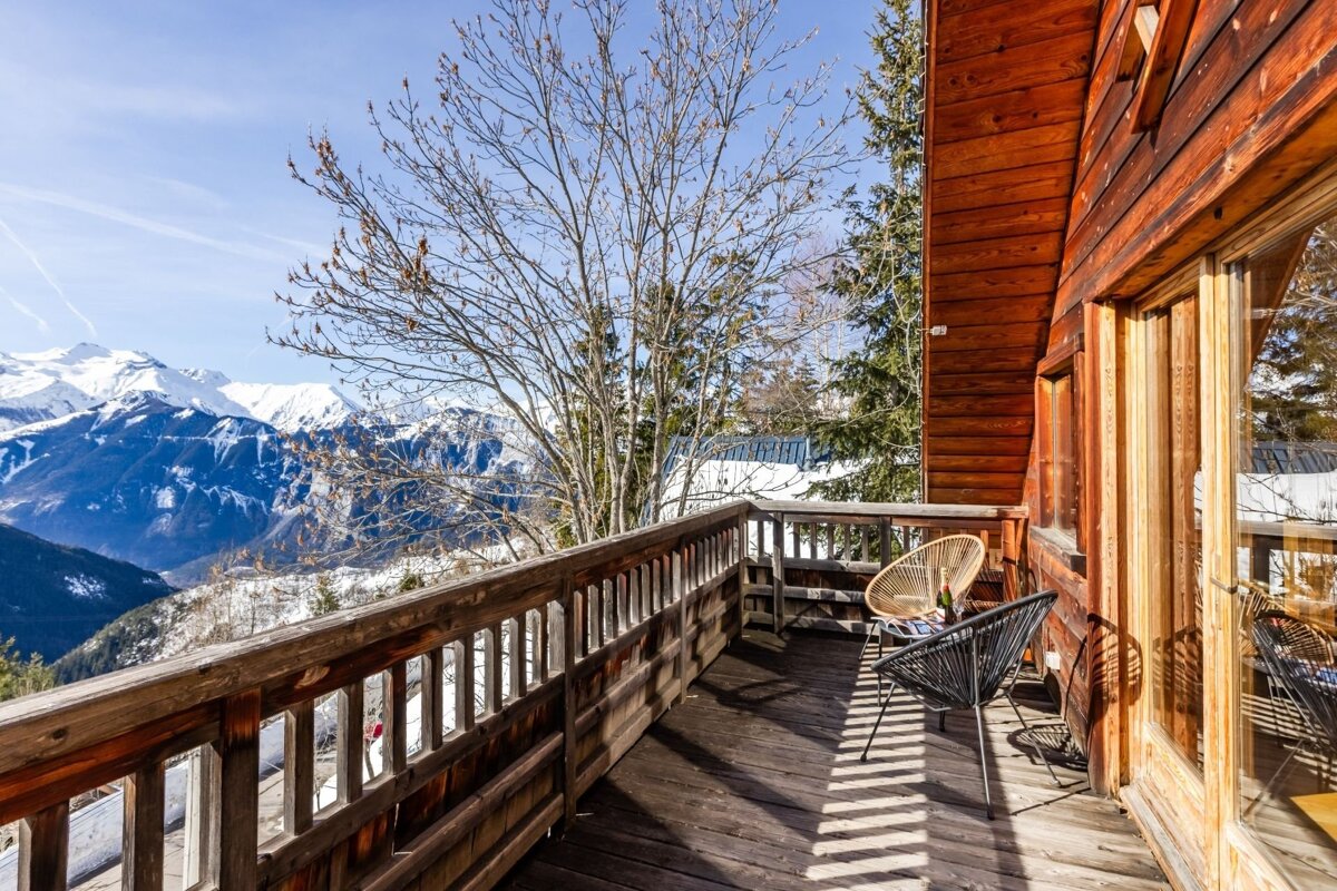 Sunny wooden chalet balcony with two chairs offering a breathtaking view of snow-capped mountain peaks.