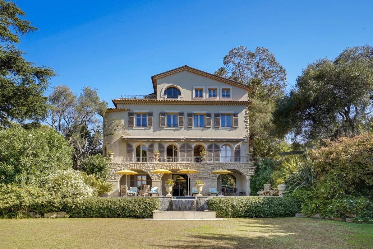 A grand Mediterranean villa with balconies, shutters, and a stone facade, surrounded by a lush green garden and trees under a clear blue sky.