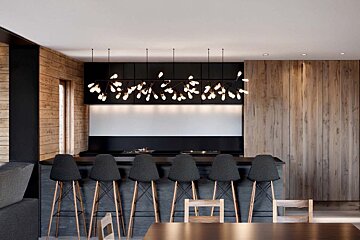 Modern kitchen bar with six dark stools, wooden paneling, and a unique branch-like chandelier with glowing lights.