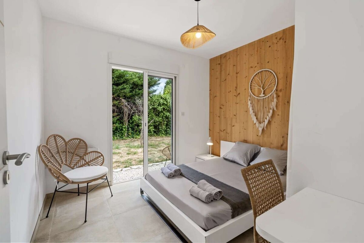 A modern bedroom with white walls, a wooden accent wall, a bed with grey linens, a rattan chair, and a sliding glass door to a garden.
