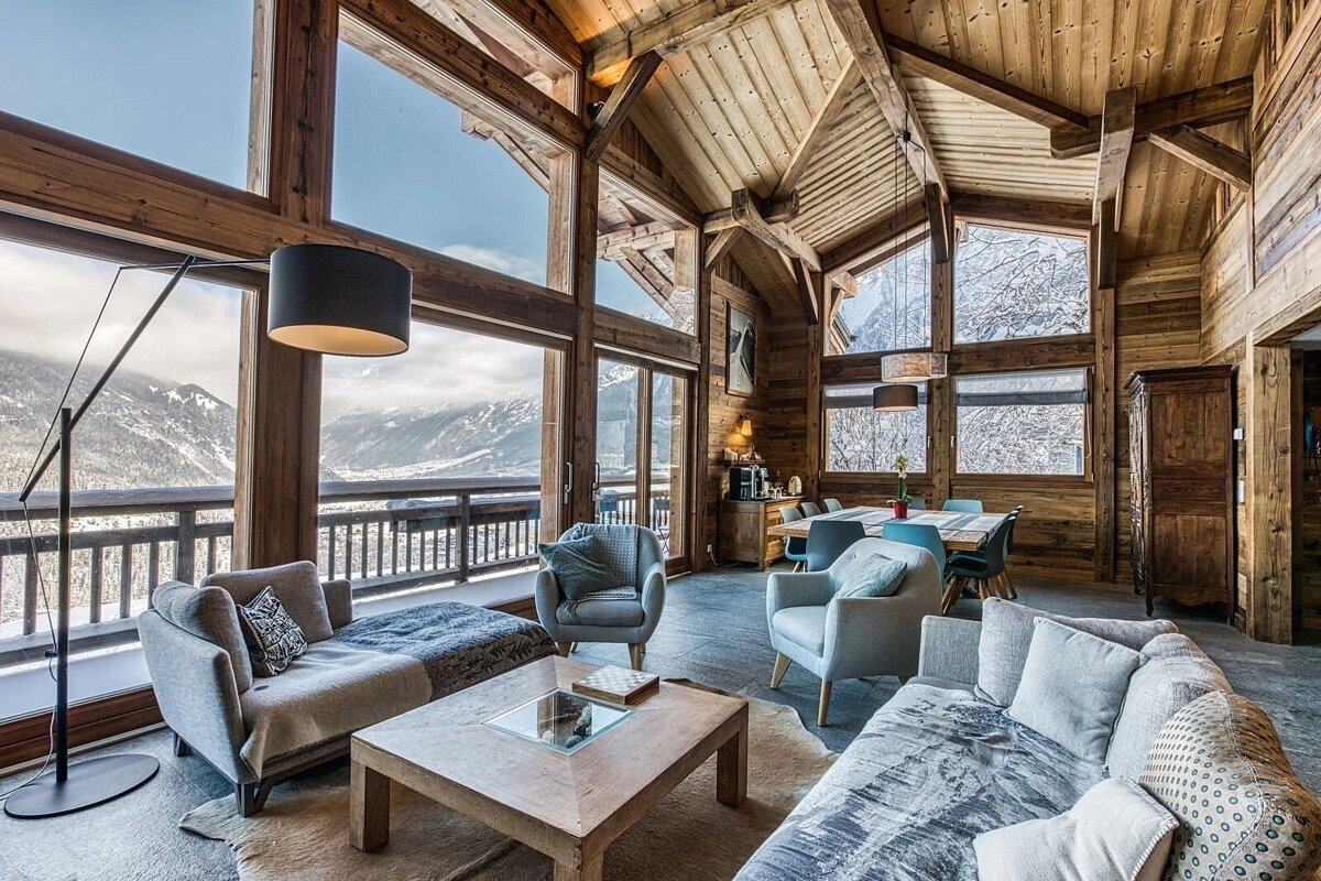 A cozy, rustic wooden chalet living room with vaulted ceilings, large windows offering stunning snowy mountain views, and a modern living and dining space.