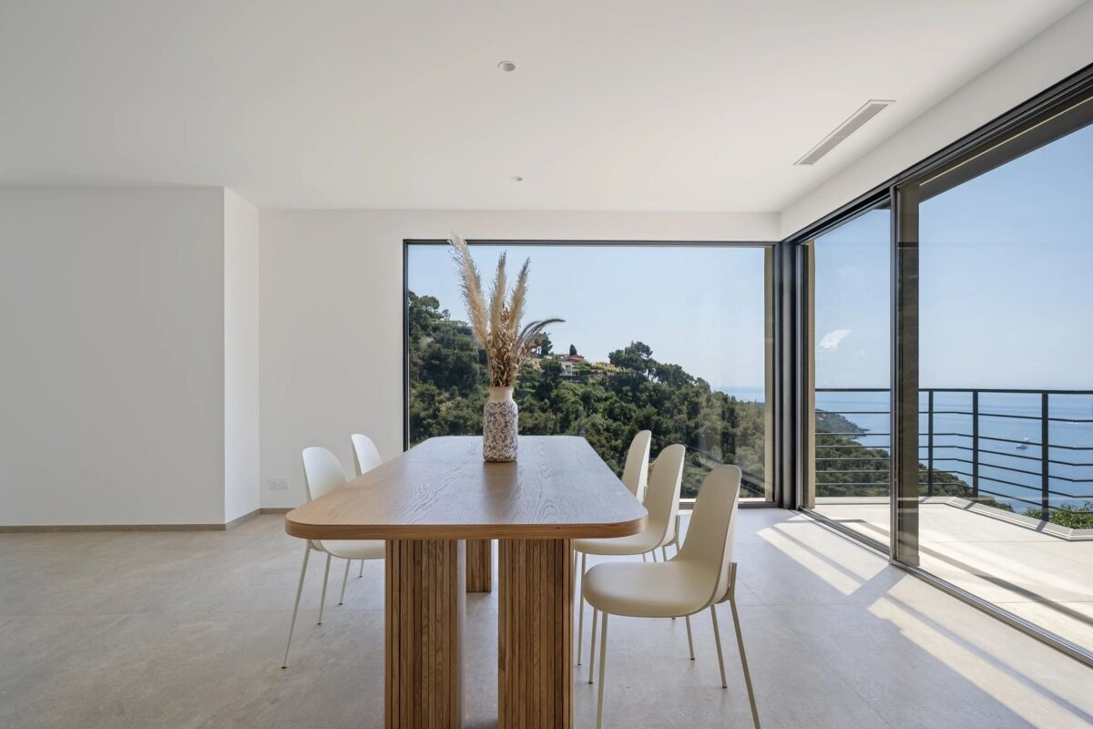 Bright, modern dining room with a wooden table, white chairs, and large windows overlooking green hills and the sea.