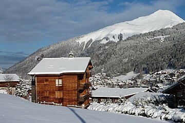 A picturesque snowy mountain village with wooden chalets under a blue sky, surrounded by snow-covered forests and majestic peaks.