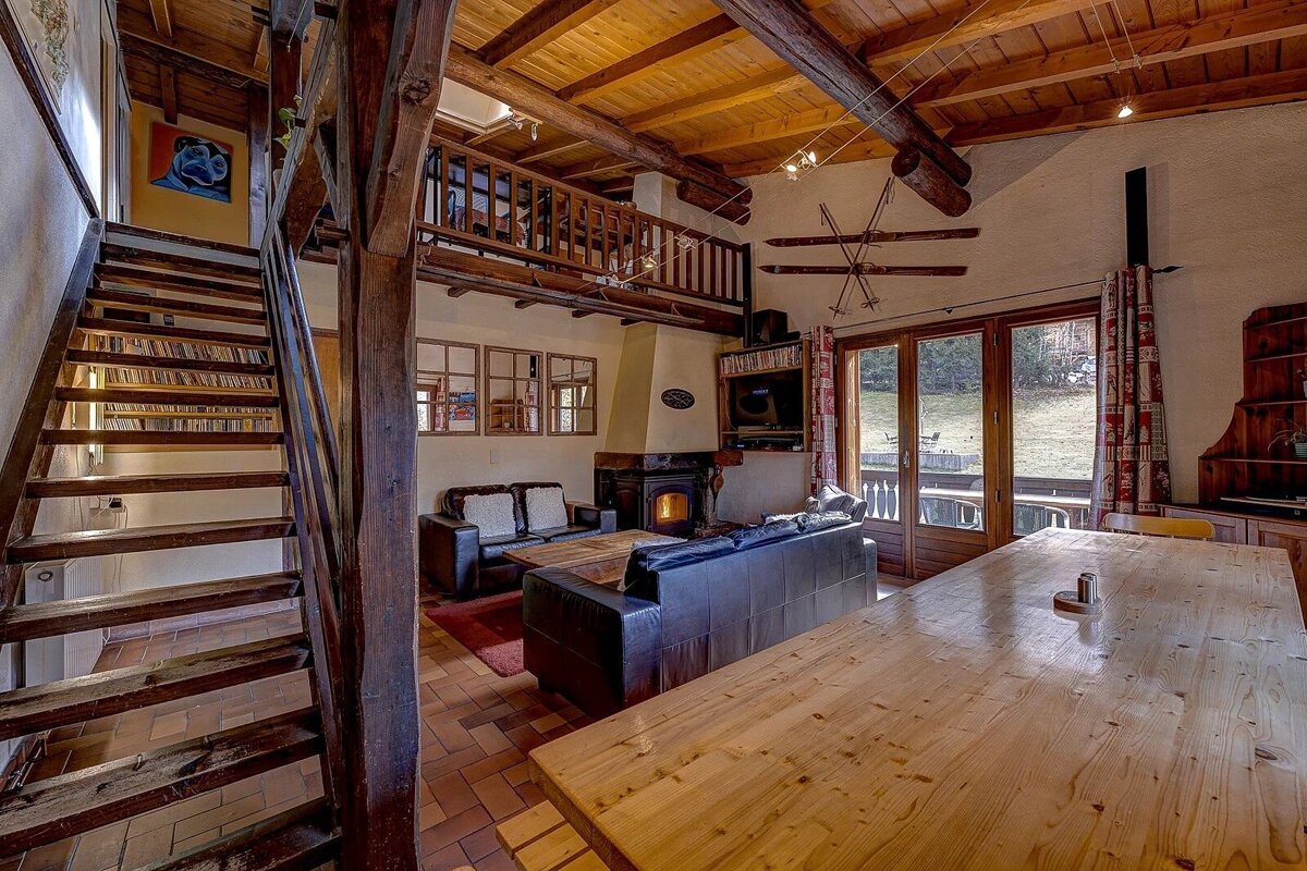 Rustic, cozy chalet living space with exposed wood beams, fireplace, leather sofas, a large dining table, and wooden stairs to a loft.