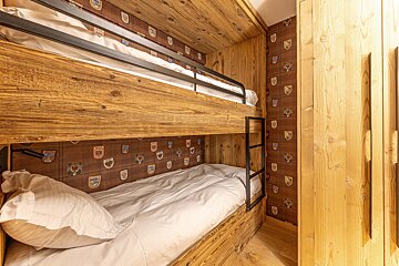 A cozy, rustic room features wooden bunk beds with white bedding, patterned crest wallpaper, and a light-toned wooden wardrobe.