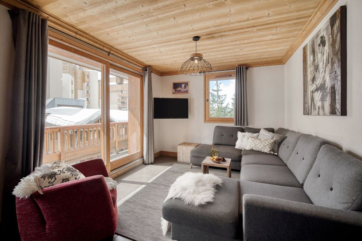 A cozy, modern chalet living room with a grey sectional sofa, wooden ceiling, TV, and a balcony view of snowy buildings.