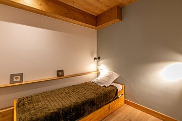 Cozy bedroom with a single wooden bed, a brown blanket, wooden ceiling beams, and soft warm lighting.