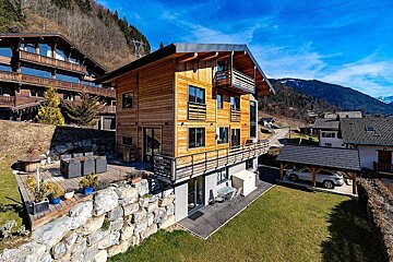 Sunny mountain scene featuring a modern wooden chalet with balconies, an outdoor patio, and other homes in the village.