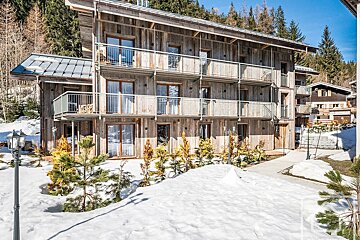 A rustic wooden chalet with multiple balconies sits snow-covered amidst a winter forest scene, under a bright sky.