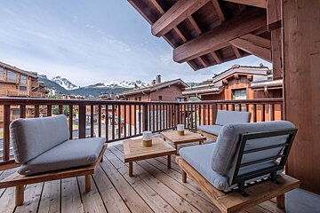 Wooden balcony with grey cushioned furniture and tables, overlooking a mountain village and snowy peaks under a clear sky.