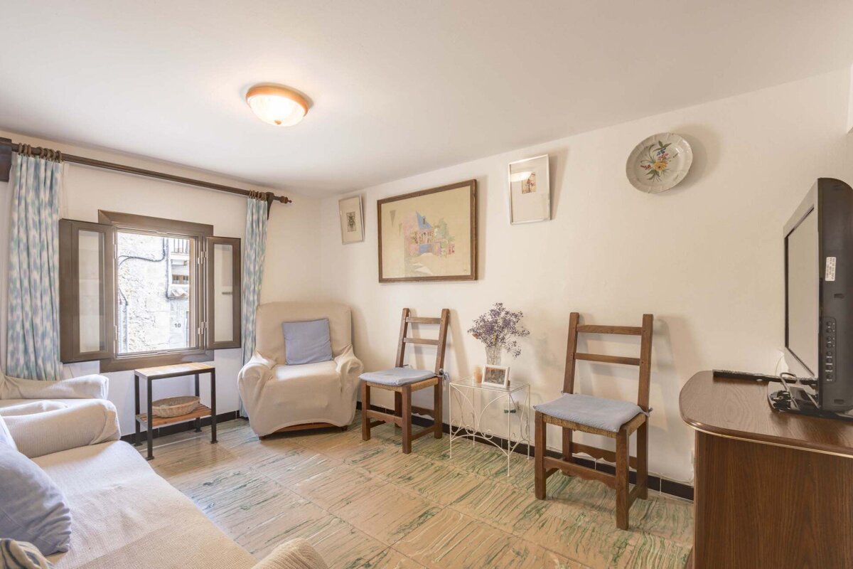 A cozy living room features a sofa, armchair, and wooden chairs. White walls display art, a window with patterned curtains, a TV, and a tiled floor.