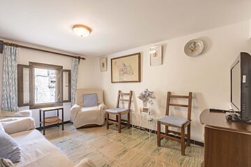 A cozy living room features a sofa, armchair, and wooden chairs. White walls display art, a window with patterned curtains, a TV, and a tiled floor.