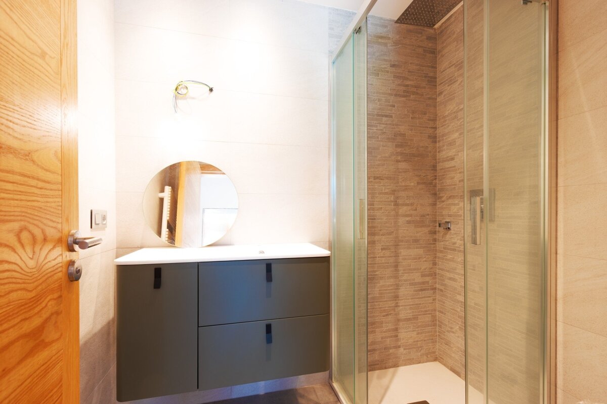 A modern bathroom featuring a sage green vanity with a round mirror, a glass shower enclosure with tiled walls, and a wooden door.