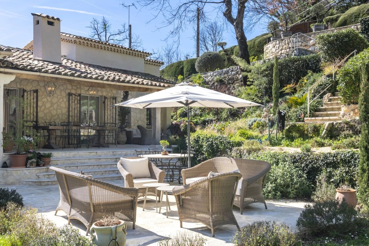 A stone house with dining and lounge patios, including wicker chairs under a white umbrella, nestled in a lush, terraced garden with steps.