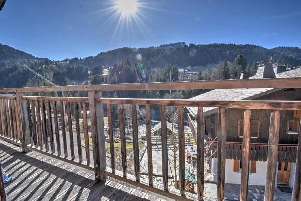 A sunny mountain view from a wooden balcony, showcasing a village, pine forests, and ski slopes under a bright blue sky.