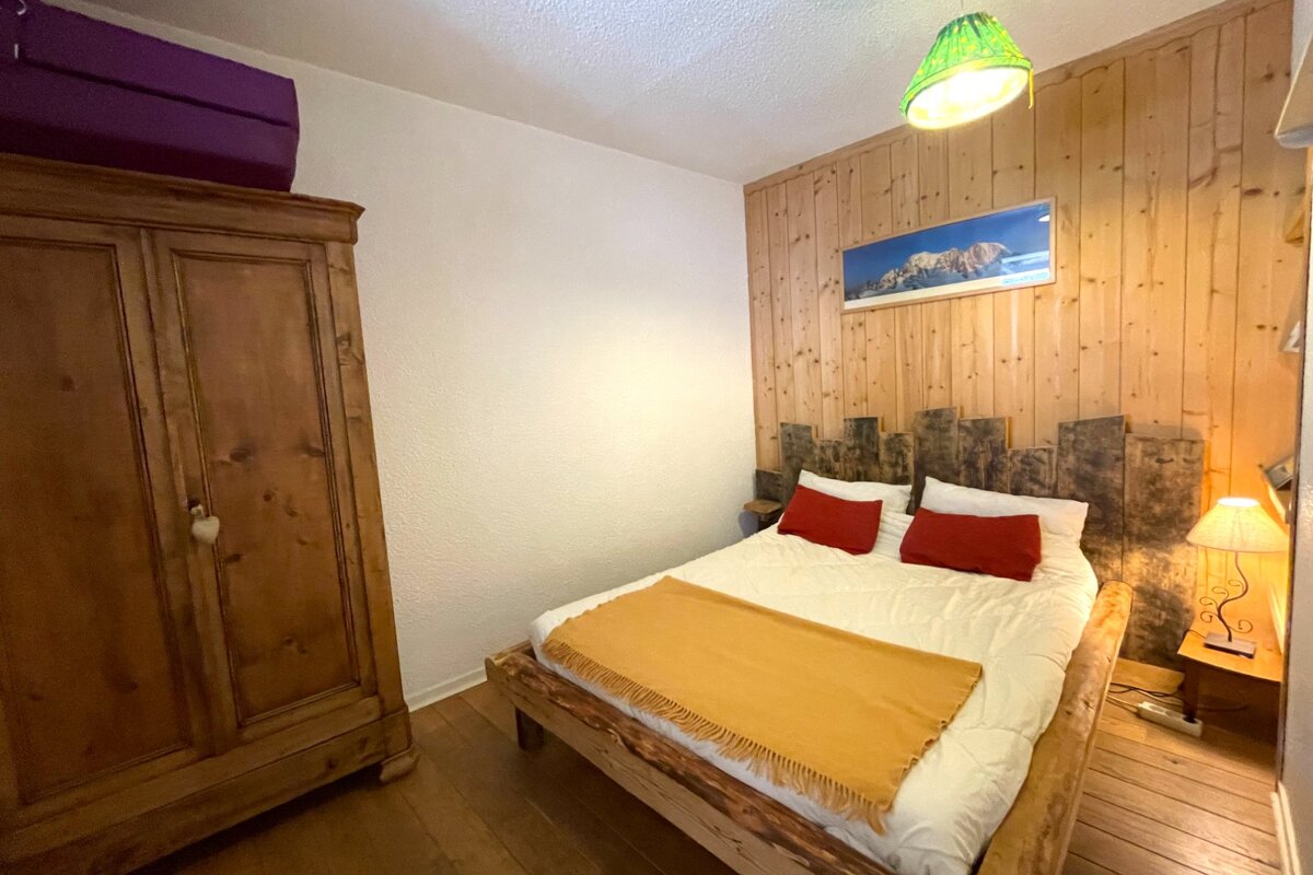 A rustic bedroom featuring a wooden bed, wardrobe, and wood-paneled wall with a mountain picture. Bed has white sheets, red pillows, yellow throw.