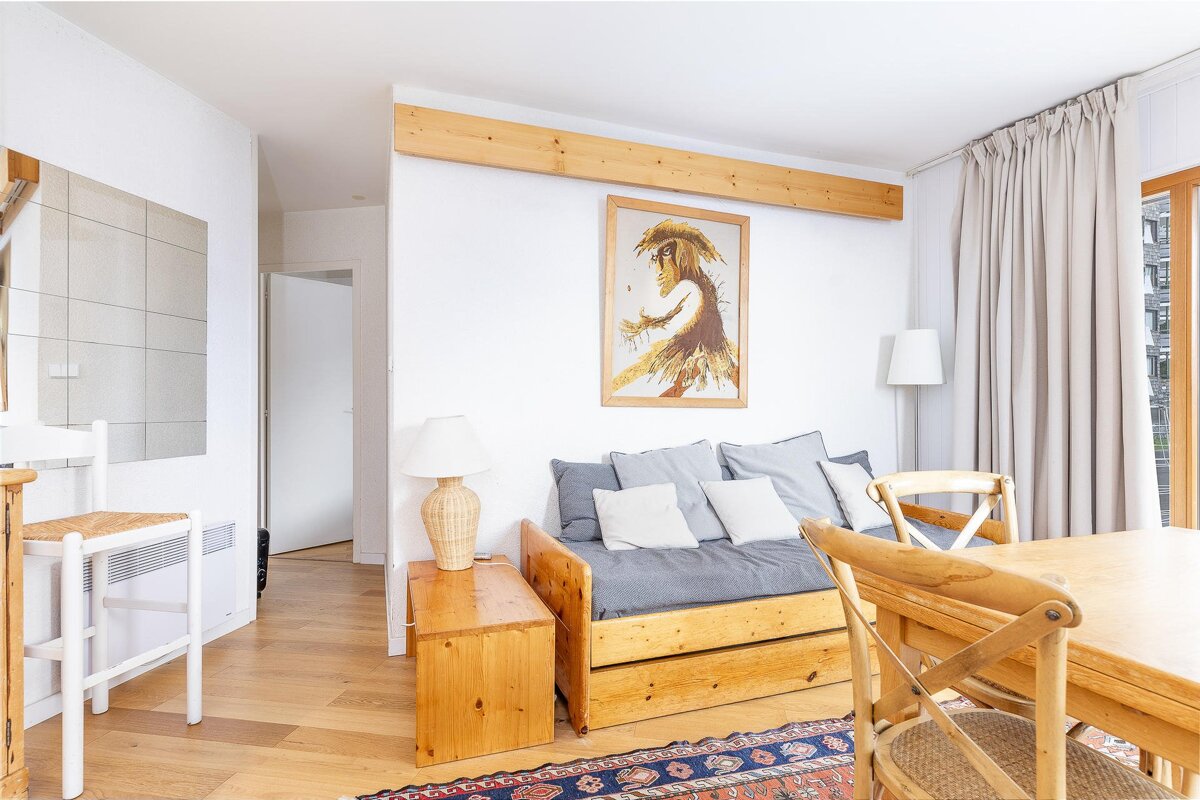 A cozy, rustic living room featuring a wooden daybed, dining table, light wood floors, a framed artwork, and beige curtains.