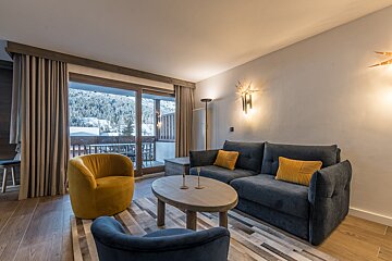 A cozy, modern living room with a blue sofa, yellow armchair, and a stunning balcony view of snowy mountains and a forest.