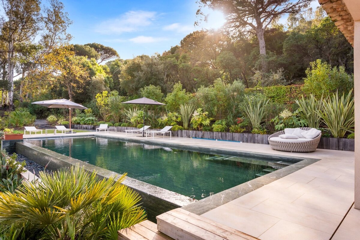 A luxurious outdoor swimming pool surrounded by lush greenery and trees, featuring lounge chairs, umbrellas, and a round daybed.