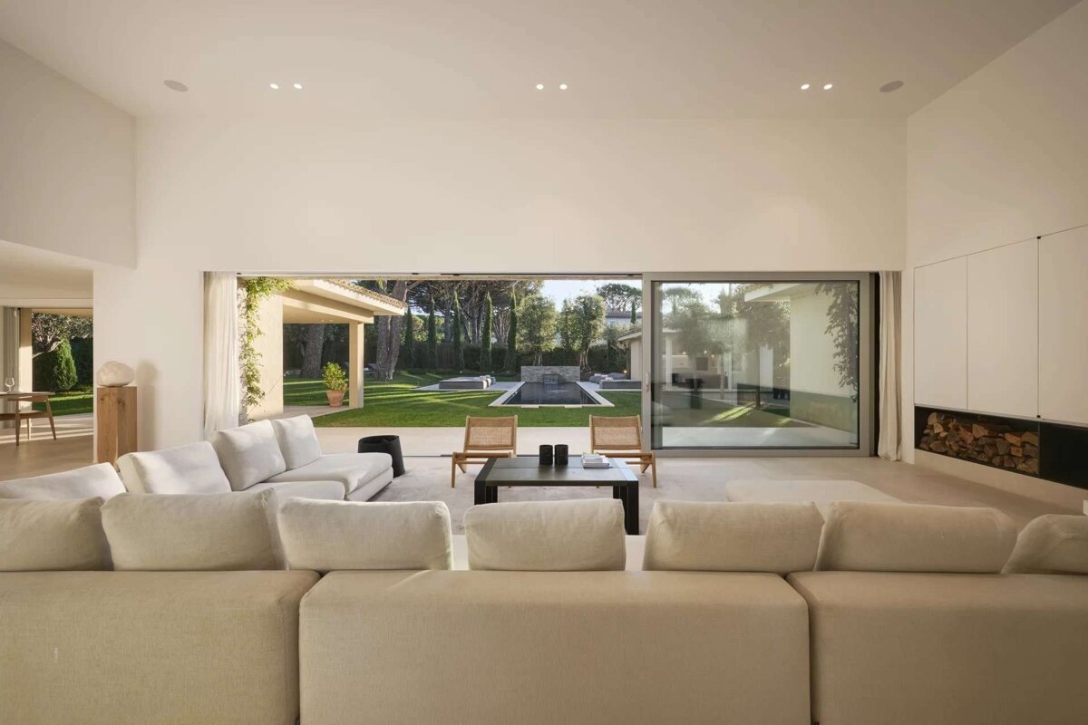 A bright, modern living room with cream sofas and expansive glass doors opening to a manicured lawn, swimming pool, and patio area.